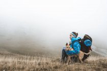 Vue latérale du voyageur détendu en veste bleu vif avec sac à dos collant chien brun et assis dans un champ sec dans la brume brumeuse en montagne — Photo de stock