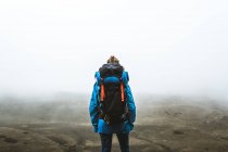 Back view of calm tranquil woman well equipped in bright blue jacket with backpack standing on top and looking at grey valley and mountains in mist — Stock Photo