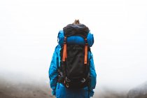 Rückenansicht einer ruhigen, gut ausgerüsteten Frau in hellblauer Jacke mit Rucksack, die oben steht und auf graue Täler und Berge im Nebel blickt — Stockfoto