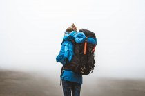 Vista lateral da mulher tranquila calma bem equipada em casaco azul brilhante com mochila em pé no topo e olhando para o vale cinza e montanhas em névoa — Fotografia de Stock