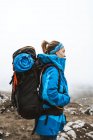 Vista lateral da mulher tranquila em casaco azul brilhante com mochila em pé na colina rochosa e olhando para longe — Fotografia de Stock