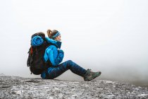 Side view of tranquil woman in bright blue jacket with backpack sitting on rocky hill and looking away — Stock Photo