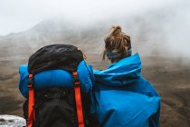 Rückansicht einer ruhigen Frau in hellblauer Jacke mit Rucksack, die auf einem felsigen Hügel sitzt und wegschaut — Stockfoto