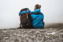 Visão traseira da mulher tranquila em casaco azul brilhante com mochila sentada na colina rochosa e olhando para longe — Fotografia de Stock