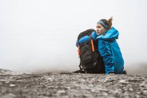 Rückansicht einer ruhigen Frau in hellblauer Jacke mit Rucksack, die auf einem felsigen Hügel sitzt und wegschaut — Stockfoto