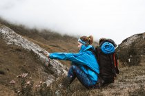 Side view of unrecognizable tranquil woman in bright blue jacket having rest sitting with backpack enjoying views looking away in dry valley in foggy haze — Stock Photo