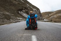 Back view of tranquil woman in bright blue jacket with backpack sitting on road and looking away — Stock Photo