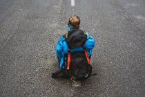 From above back view of tranquil woman in bright blue jacket with backpack sitting on road and looking away — Stock Photo