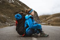 Side view of tranquil woman in bright blue jacket with backpack sitting on road and looking away — Stock Photo