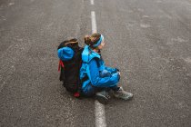 Зверху спокійна жінка в яскраво-блакитній куртці з рюкзаком сидить на дорозі і дивиться в сторону — стокове фото