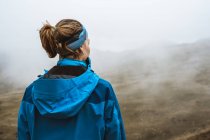Vista posterior de la mujer tranquila en chaqueta azul brillante con mochila de pie en la colina rocosa y mirando hacia otro lado - foto de stock
