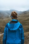 Вид ззаду спокійна жінка в яскраво-блакитній куртці з рюкзаком, що стоїть на скелястому пагорбі і дивиться вбік — стокове фото