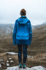 Back view of tranquil woman in bright blue jacket with backpack standing on rocky hill and looking away — Stock Photo