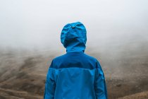 Rückansicht einer ruhigen Frau in hellblauer Jacke mit Rucksack, die auf einem felsigen Hügel steht und wegschaut — Stockfoto