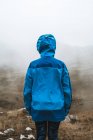Vista posterior de la mujer tranquila en chaqueta azul brillante con mochila de pie en la colina rocosa y mirando hacia otro lado - foto de stock
