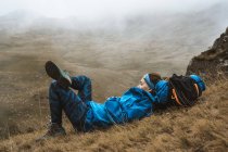 Вид збоку спокійна жінка в яскраво-блакитній куртці, спираючись на рюкзак і лежачи з схрещеною ногою в сухій долині в туманному туманному тумані — стокове фото