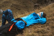 Vista lateral da mulher tranquila em casaco azul brilhante com descanso apoiado na mochila e deitado com a perna cruzada no vale seco na névoa nebulosa — Fotografia de Stock