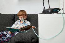 Menino em máscara de oxigênio usando nebulizador durante a inalação enquanto sentado no sofá e livro de leitura — Fotografia de Stock