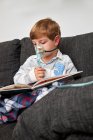 Menino em máscara de oxigênio usando nebulizador durante a inalação enquanto sentado no sofá e livro de leitura — Fotografia de Stock