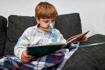 Garçon en masque à oxygène en utilisant un nébuliseur pendant l'inhalation tout en étant assis sur le canapé et le livre de lecture — Photo de stock