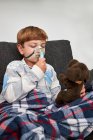Criança alegre respirando máscara de oxigênio durante a inalação e brincando com brinquedo macio no sofá em casa — Fotografia de Stock