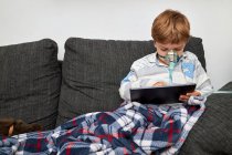 Ragazzo malato in maschera di ossigeno seduto sul divano a casa e giocare al videogioco su tablet — Foto stock