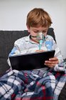 Garçon malade en masque à oxygène assis sur le canapé à la maison et jouer à un jeu vidéo sur tablette — Photo de stock