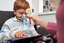Crop unrecognizable loving mother with infrared thermometer measuring temperature of boy breathing in oxygen mask during inhalation procedure and watching cartoon on tablet at home — Stock Photo