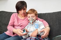 Mulher carinhosa usando oxímetro de pulso no dedo do menino doente em máscara de oxigênio usando nebulizador durante a inalação em casa — Fotografia de Stock