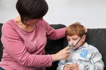 Content mother putting on oxygen mask on face of son for inhalation procedure while sitting on sofa at home — Stock Photo
