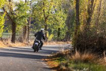 Visão traseira da pessoa irreconhecível em jaqueta de couro e capacete andar de bicicleta na estrada de asfalto no dia ensolarado de outono no campo — Fotografia de Stock