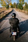 Vista posteriore di persona irriconoscibile in giacca di pelle e casco in bicicletta su strada asfaltata in soleggiata giornata autunnale in campagna — Foto stock