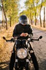 Concentrated aged male racer in leather jacket fastening helmet and sitting on motorbike in autumn sunny day — Stock Photo