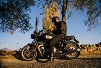 Racer uomo invecchiato concentrato in giacca di pelle casco di fissaggio e seduto sulla moto in autunno giornata di sole — Foto stock