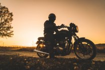 Motociclista sem rosto no capacete de equitação motocicleta à noite pôr do sol na estrada rural — Fotografia de Stock