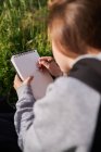 Back view of unrecognizable teen girl hiding face while sitting in meadow and drawing in sketchbook enjoying sunny day in countryside — Stock Photo