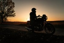 Motociclista sin rostro en casco a caballo motocicleta en la puesta del sol por la noche en la carretera rural - foto de stock