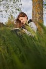 Encantada menina adolescente sentado no prado e desenho em caderno de esboços enquanto desfruta de dia ensolarado no campo encostado no tronco da árvore — Fotografia de Stock