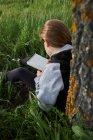 Back view of unrecognizable teen girl sitting in meadow and drawing in sketchbook while enjoying sunny day in countryside leaning on tree trunk — Stock Photo