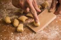 Unrecognizable cook hands pressing a piece of dough to ribbed board while preparing gnocchi on wooden table covered with flour — Stock Photo