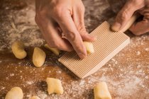 Unrecognizable cook hands pressing a piece of dough to ribbed board while preparing gnocchi on wooden table covered with flour — Stock Photo