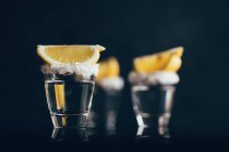 Tequila tiros com sal e limão colocados na superfície reflexiva contra fundo escuro — Fotografia de Stock