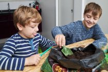 Kind mit Schaufel nimmt Erde aus Plastiktüte am Tisch gegen lächelndes Geschwisterchen mit Ökobecher im Haus — Stockfoto