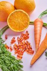 Зверху композиція розкиданих вітамінних таблеток розташована на рожевому столі біля стиглої моркви і соковитих апельсинів — стокове фото