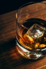 Du dessus tasse en verre avec du whisky froid et cube de glace placé sur la table de bois dans la pièce sombre — Photo de stock