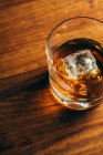 From above glass cup with cold whiskey and cube of ice placed on lumber table in dark room — Stock Photo
