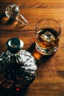 Glass of cold whiskey with ice placed on wooden table near decanter on black background — Stock Photo