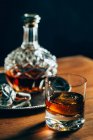 Verre de whisky froid avec glace placé sur une table en bois près du décanteur sur fond noir — Photo de stock
