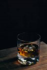 Сверху стеклянная чашка с холодным виски и кубиком льда, помещенная на деревянный стол в темной комнате — стоковое фото