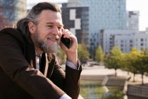 Side view of smiling bearded mature male with gray hair wearing classy suit discussing business issues during phone conversation while standing against modern city buildings — Stock Photo